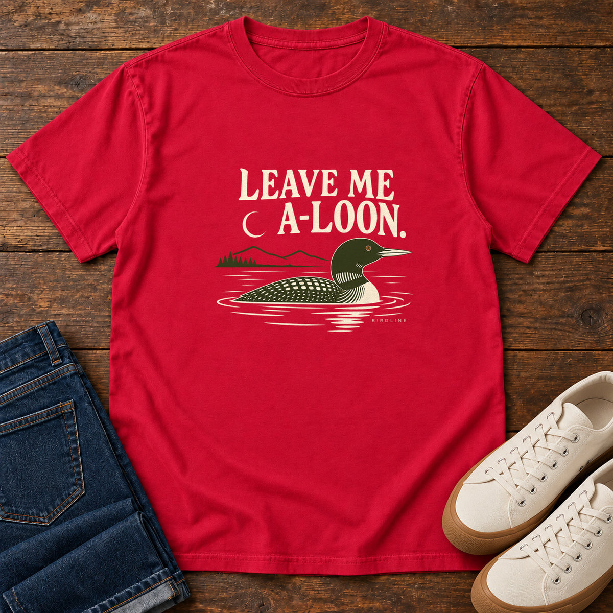 Red Leave Me A Loon T-Shirt with a green-and-cream waterbird floating below a crescent moon and cream lettering, front flat lay on wood.
