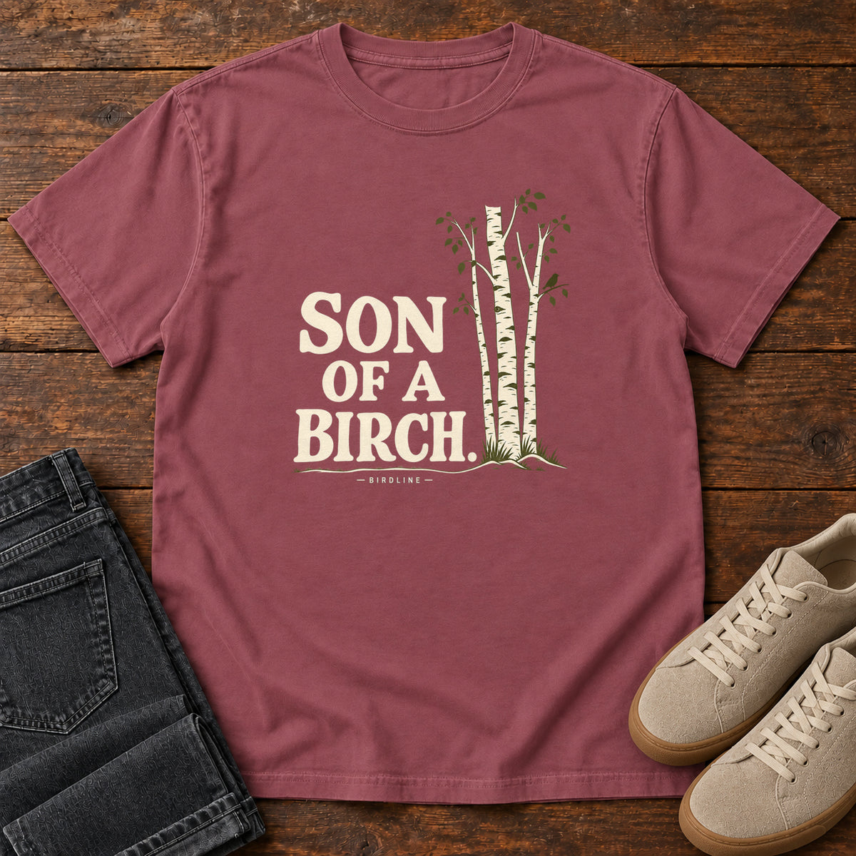 Brick Son of a Birch T-Shirt with cream lettering beside slender white-barked trees, front flat lay on wood.