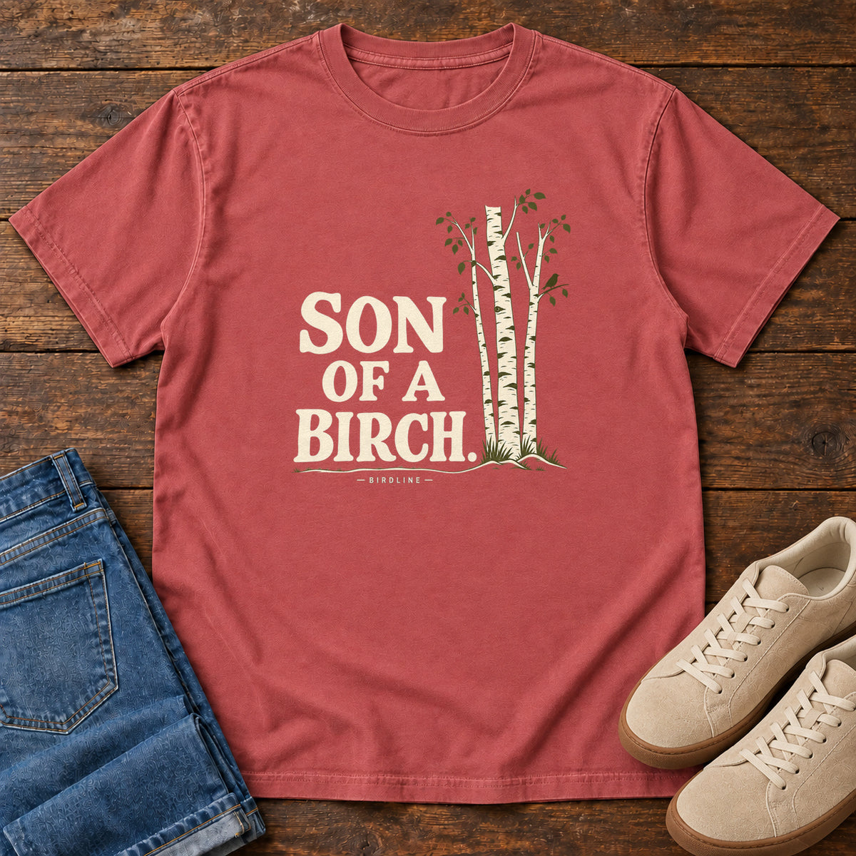 Crimson Son of a Birch T-Shirt with cream lettering beside slender white-barked trees, front flat lay on wood.