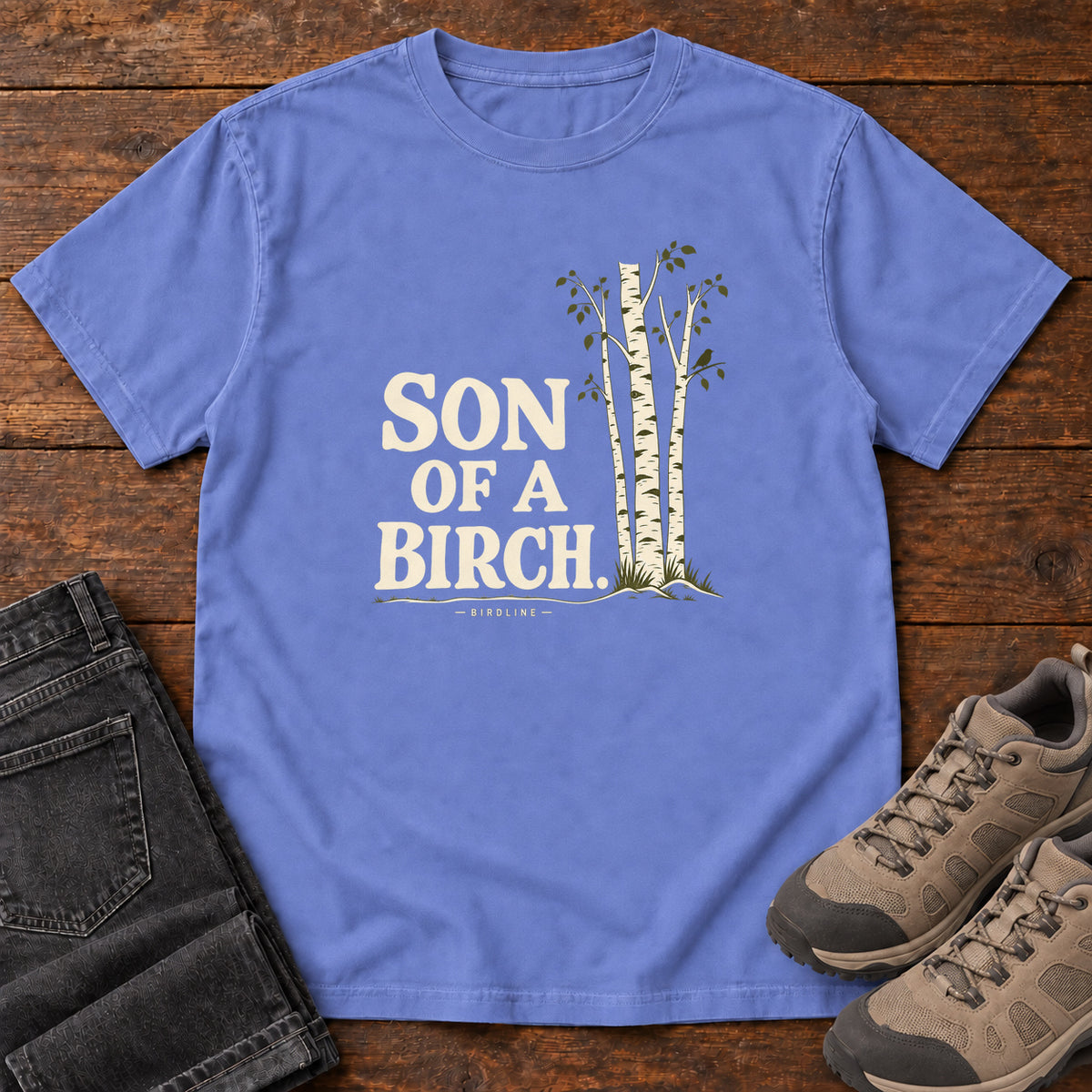 Flo Blue Son of a Birch T-Shirt with cream lettering beside slender white-barked trees, front flat lay on wood.
