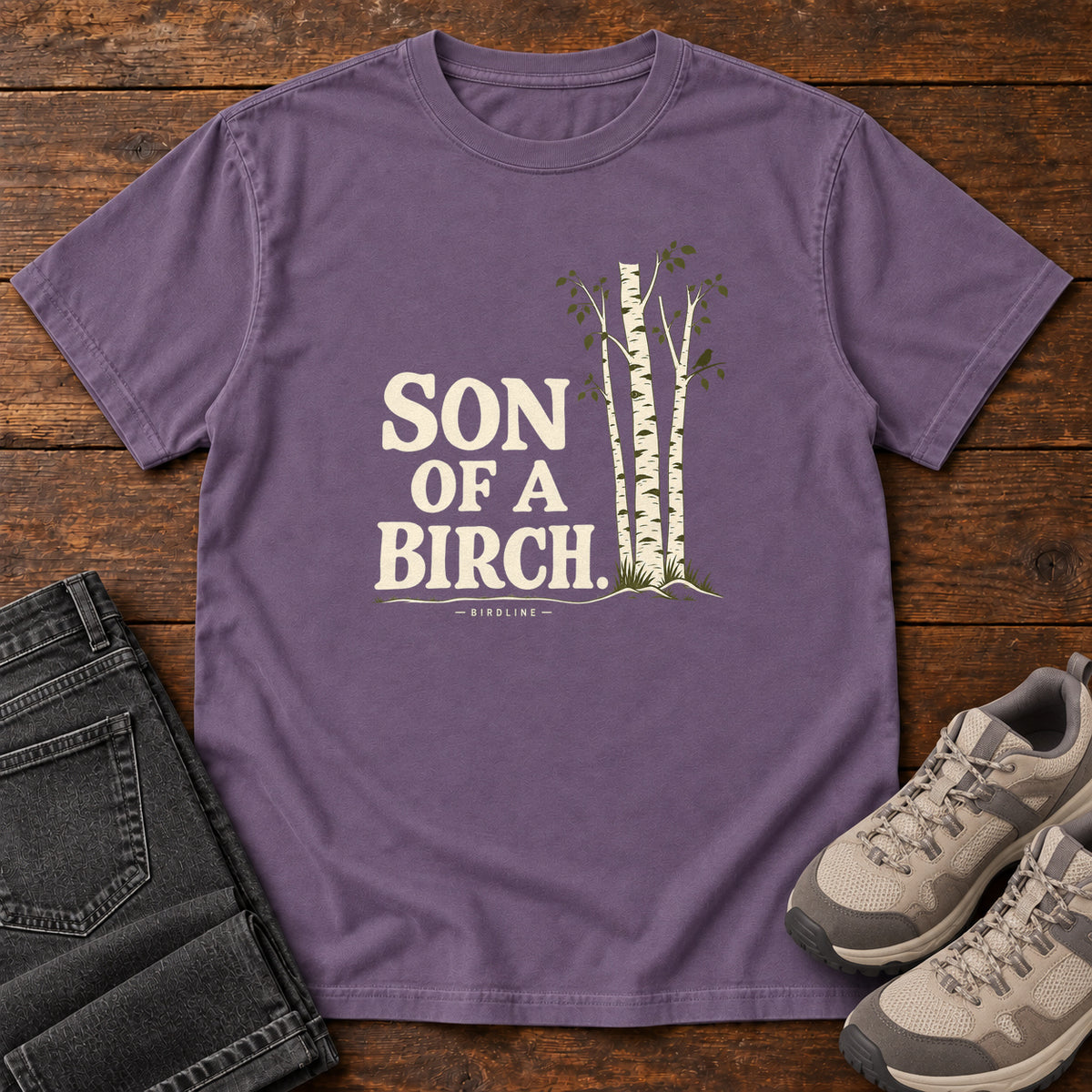 Grape Son of a Birch T-Shirt with cream lettering beside slender white-barked trees, front flat lay on wood.