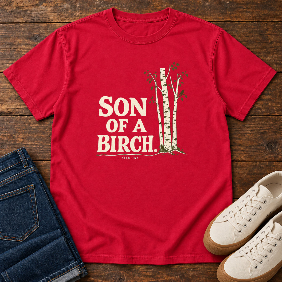 Red Son of a Birch T-Shirt with cream lettering beside slender white-barked trees, front flat lay on wood.