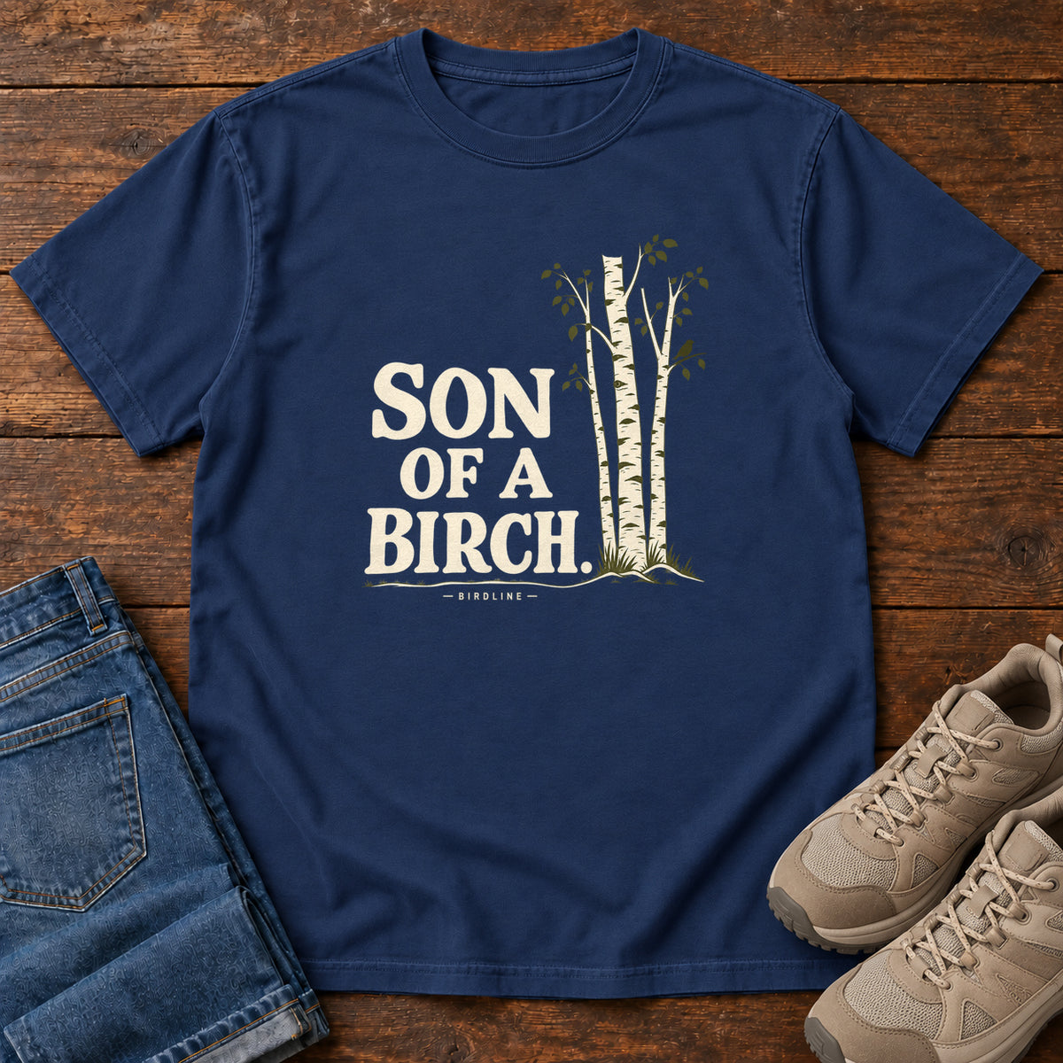 True Navy Son of a Birch T-Shirt with cream lettering beside slender white-barked trees, front flat lay on wood.