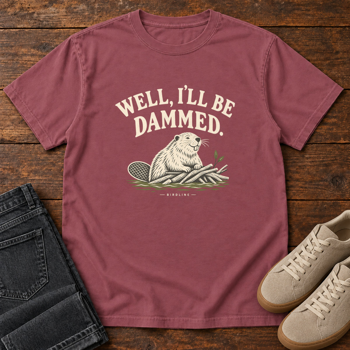 Well I'll Be Dammed T-Shirt in Brick, front laid flat on wood, featuring a cream beaver sitting beside branches beneath curved slogan lettering.