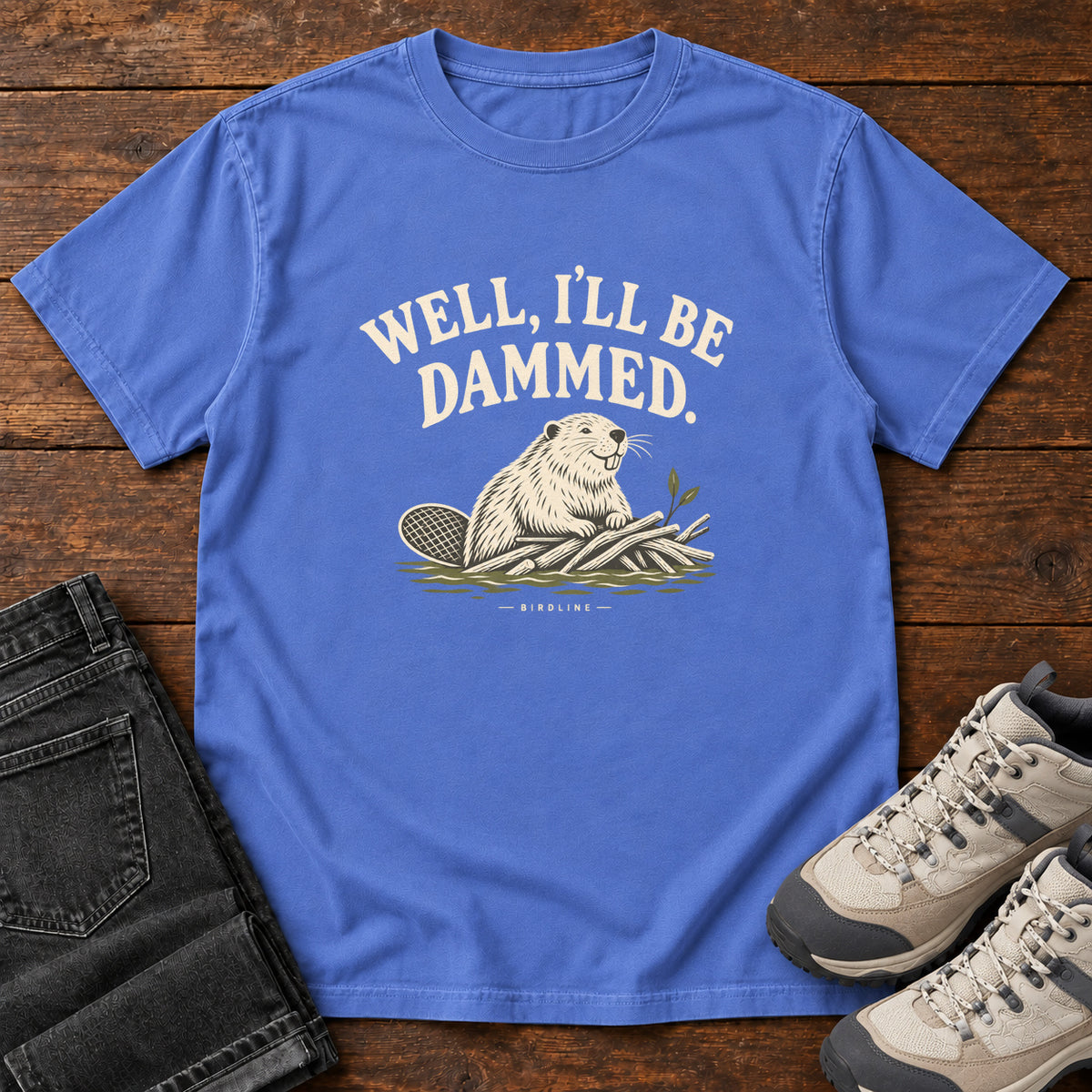 Well I'll Be Dammed T-Shirt in Mystic Blue, front laid flat on wood, featuring a cream beaver sitting beside branches beneath curved slogan lettering.