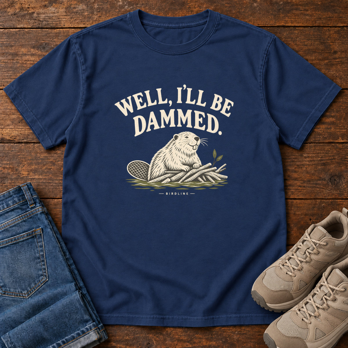 Well I'll Be Dammed T-Shirt in True Navy, front laid flat on wood, featuring a cream beaver sitting beside branches beneath curved slogan lettering.