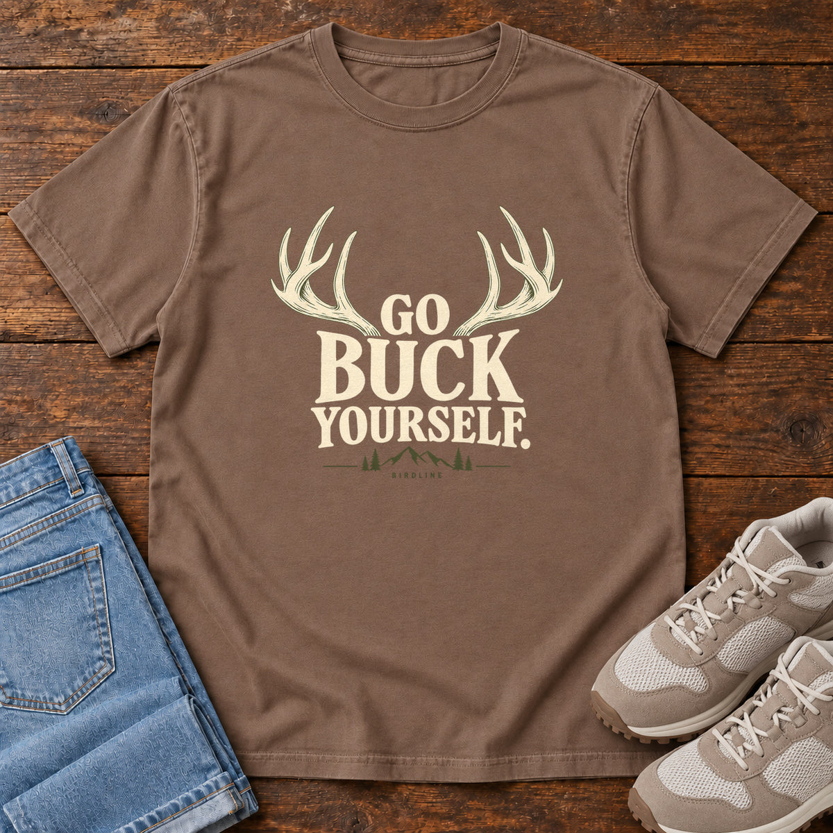 Espresso Go Buck Yourself T-Shirt with cream deer antlers framing Go Buck Yourself lettering, front flat lay on wood.
