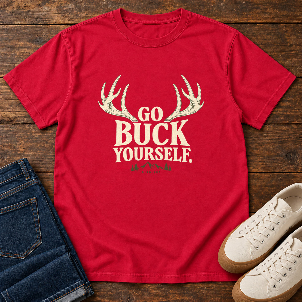 Red Go Buck Yourself T-Shirt with cream deer antlers framing Go Buck Yourself lettering, front flat lay on wood.