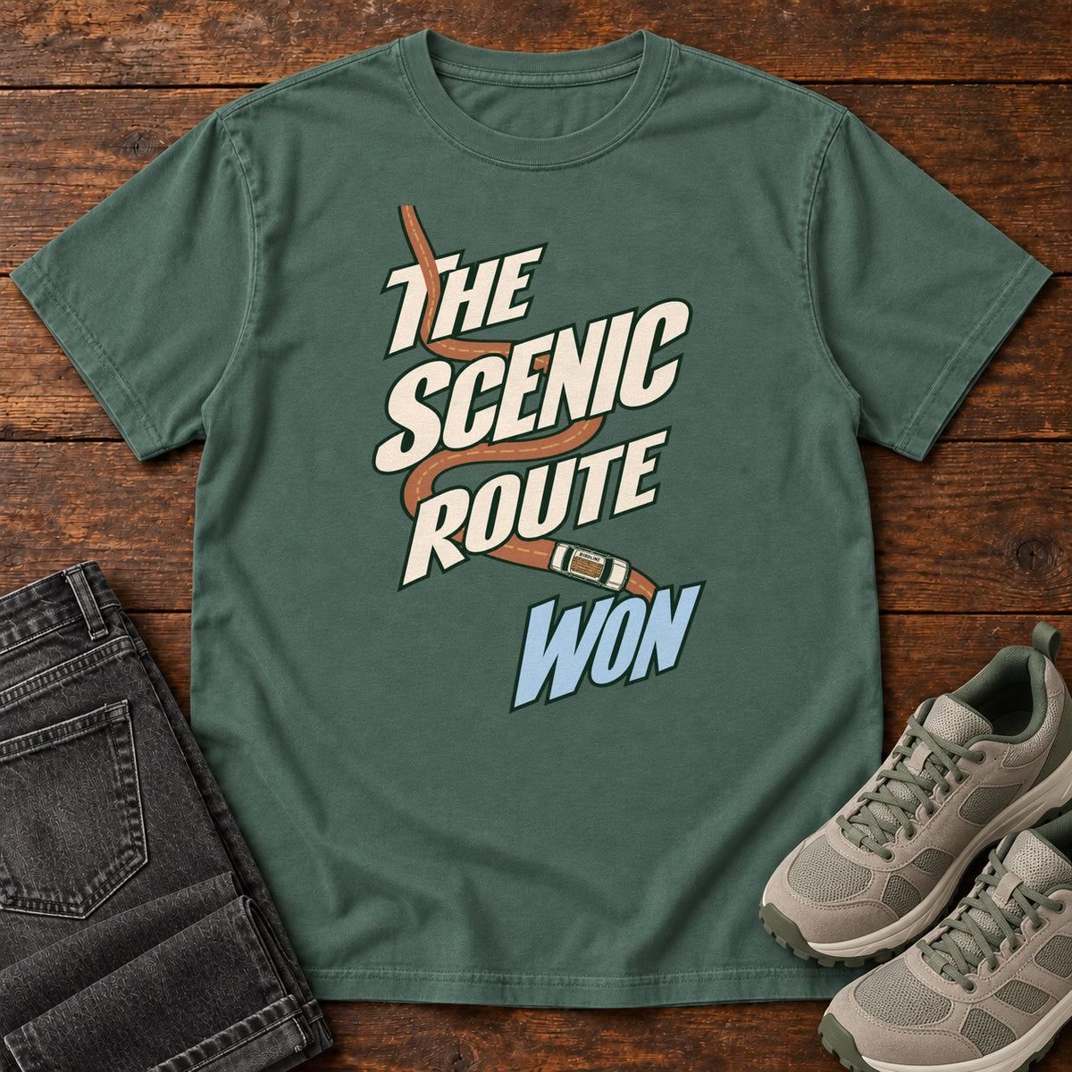 The Scenic Route Won T-Shirt in Blue Spruce, front laid flat on wood, featuring a winding orange road and tiny car behind slanted cream-and-blue slogan lettering.