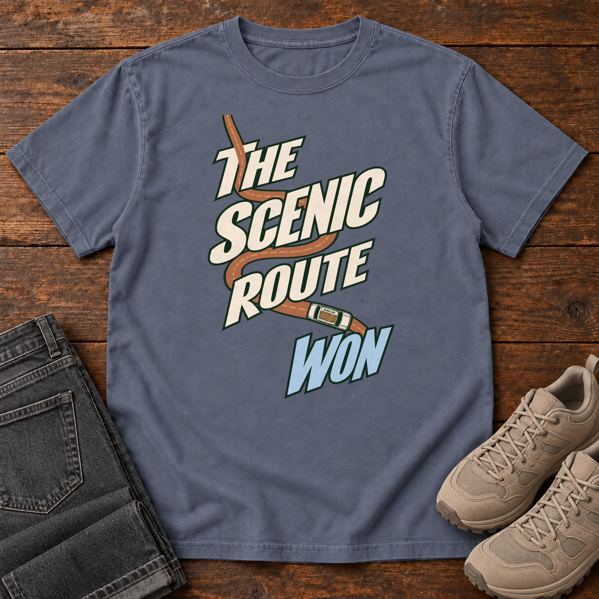 The Scenic Route Won T-Shirt in Denim, front laid flat on wood, featuring a winding orange road and tiny car behind slanted cream-and-blue slogan lettering.