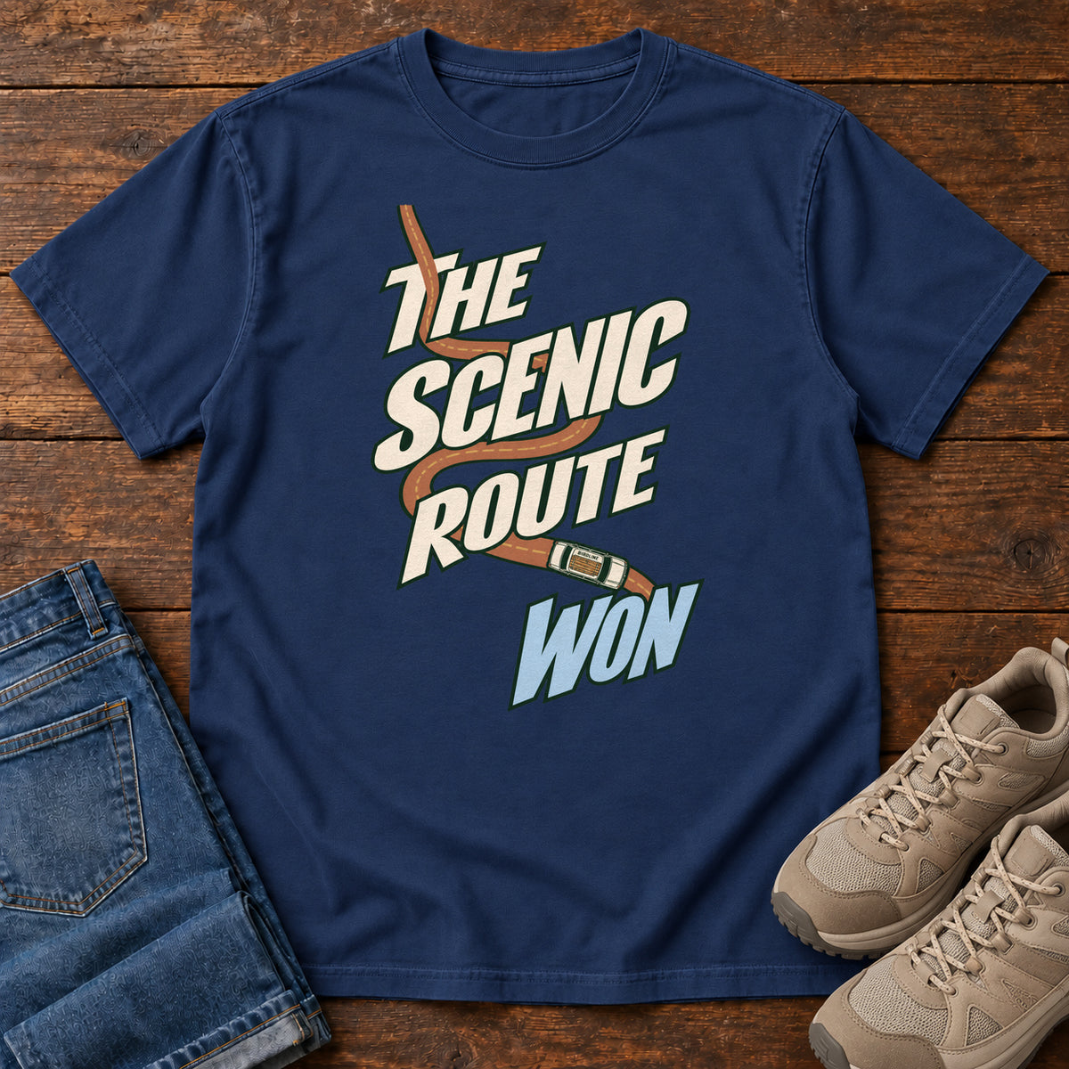 The Scenic Route Won T-Shirt in True Navy, front laid flat on wood, featuring a winding orange road and tiny car behind slanted cream-and-blue slogan lettering.
