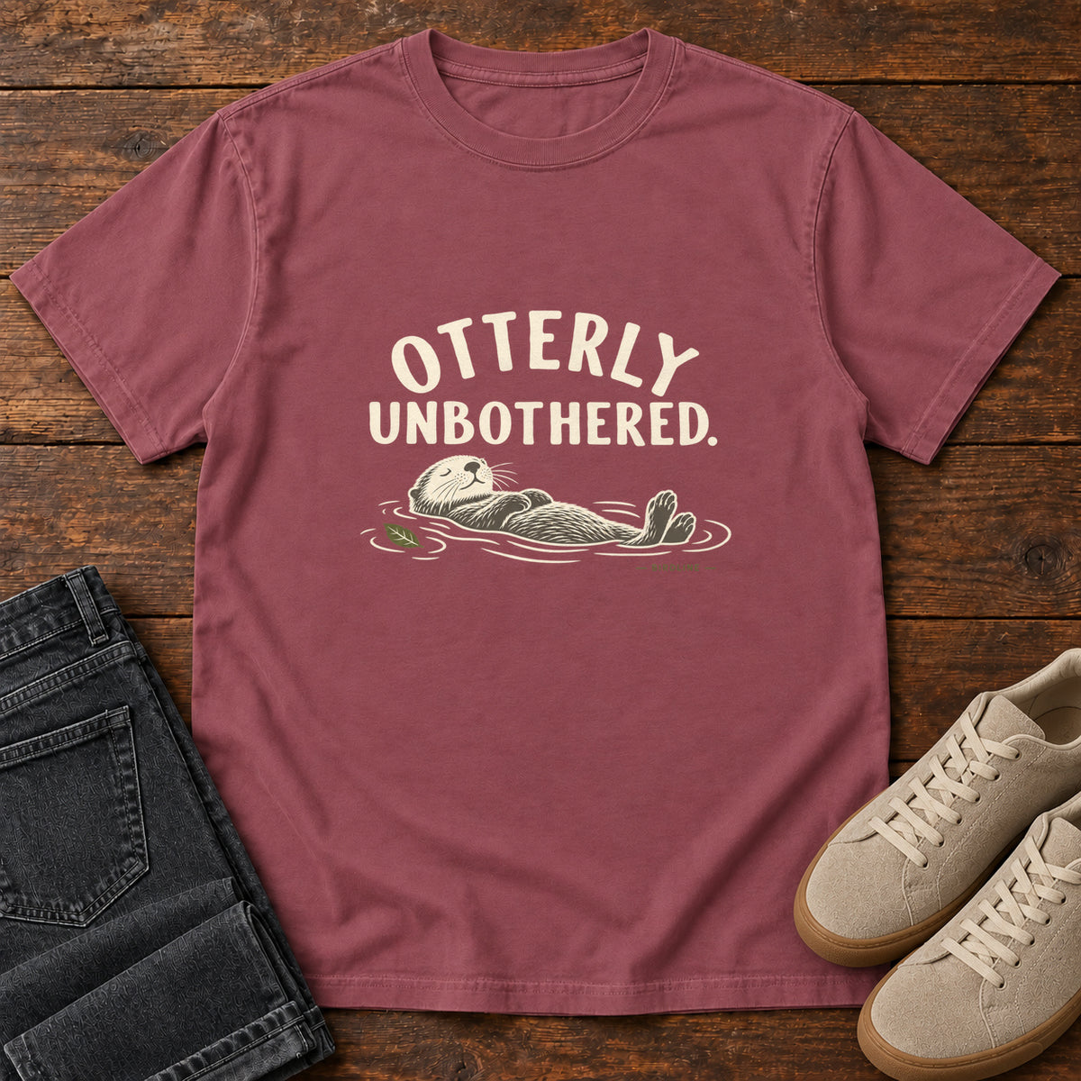 Brick Otterly Unbothered T-Shirt with a cream line drawing of an otter floating on its back beneath curved lettering; front flat lay on wood.