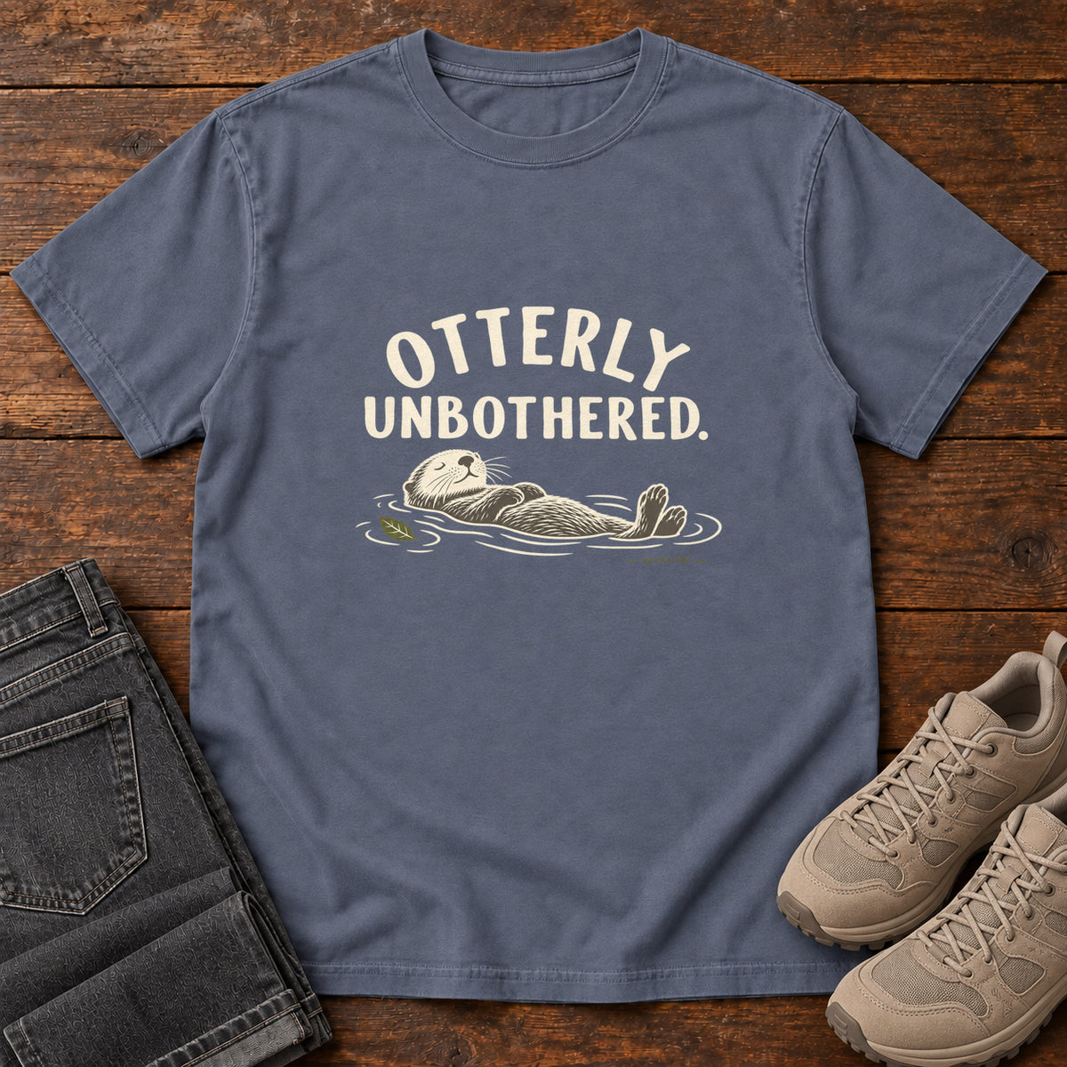 Denim Otterly Unbothered T-Shirt with a cream line drawing of an otter floating on its back beneath curved lettering; front flat lay on wood.