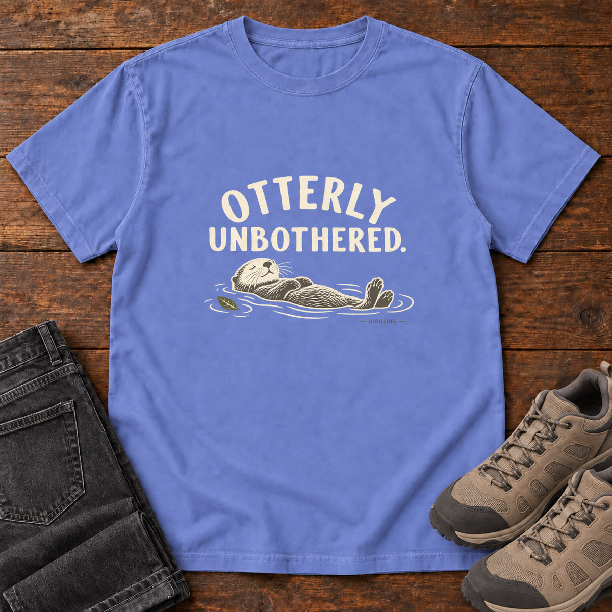 Flo Blue Otterly Unbothered T-Shirt with a cream line drawing of an otter floating on its back beneath curved lettering; front flat lay on wood.