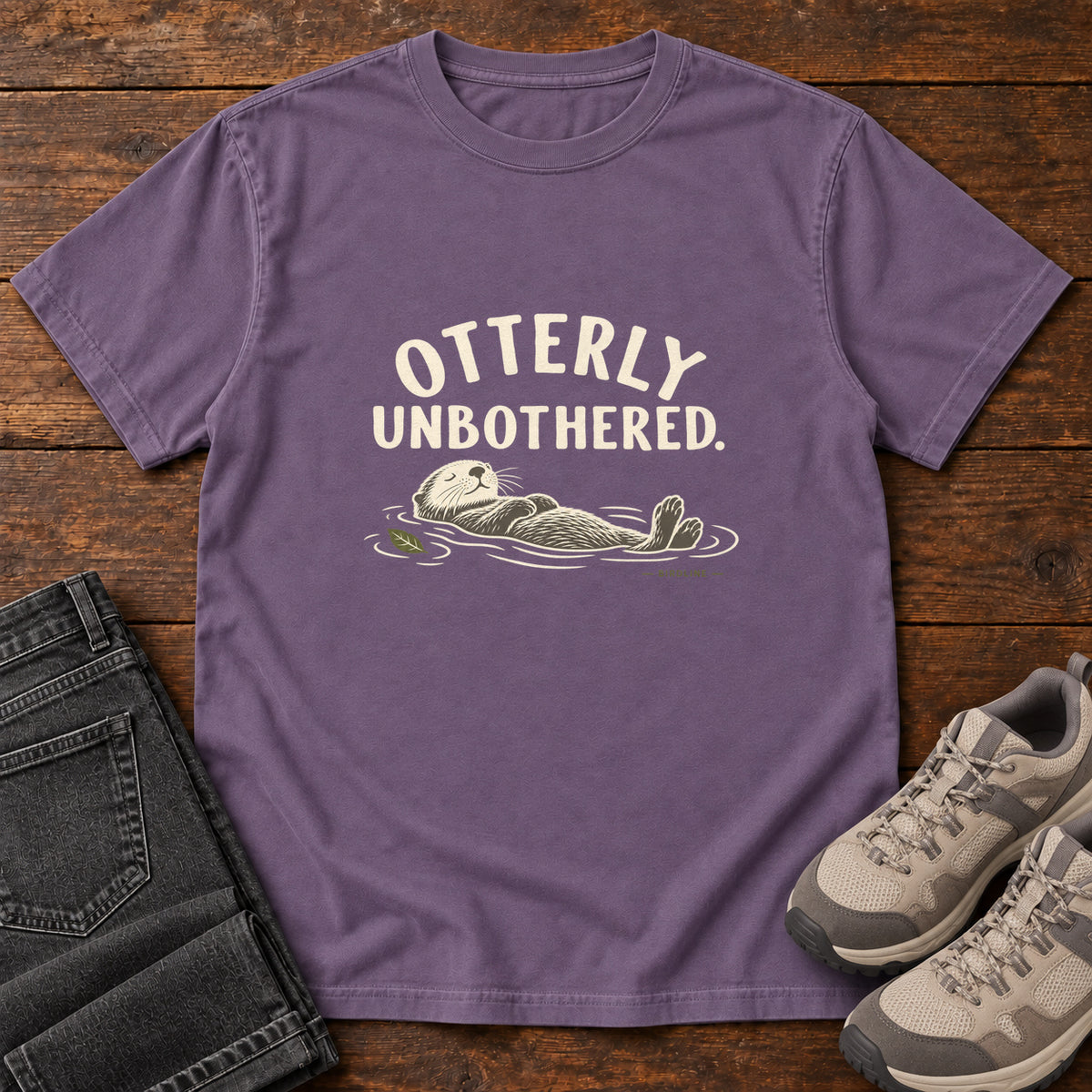 Grape Otterly Unbothered T-Shirt with a cream line drawing of an otter floating on its back beneath curved lettering; front flat lay on wood.