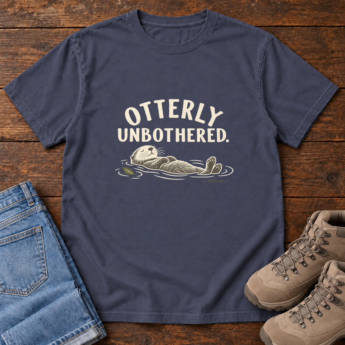 Navy Otterly Unbothered T-Shirt with a cream line drawing of an otter floating on its back beneath curved lettering; front flat lay on wood.