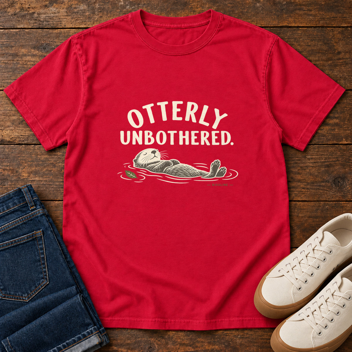 Red Otterly Unbothered T-Shirt with a cream line drawing of an otter floating on its back beneath curved lettering; front flat lay on wood.