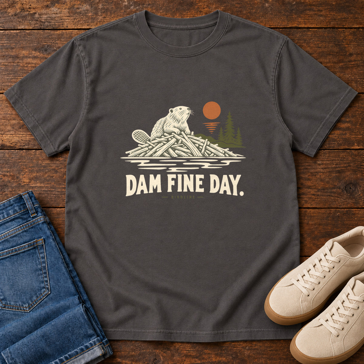 Graphite Dam Fine Day T-Shirt with a beaver on a pile of branches beside evergreens and an orange sun; front flat lay on wood.