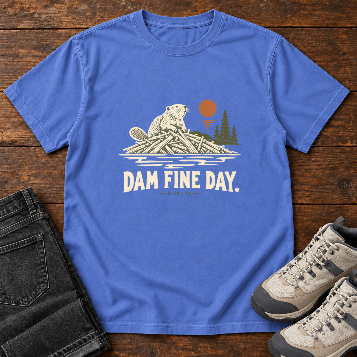Mystic Blue Dam Fine Day T-Shirt with a beaver on a pile of branches beside evergreens and an orange sun; front flat lay on wood.