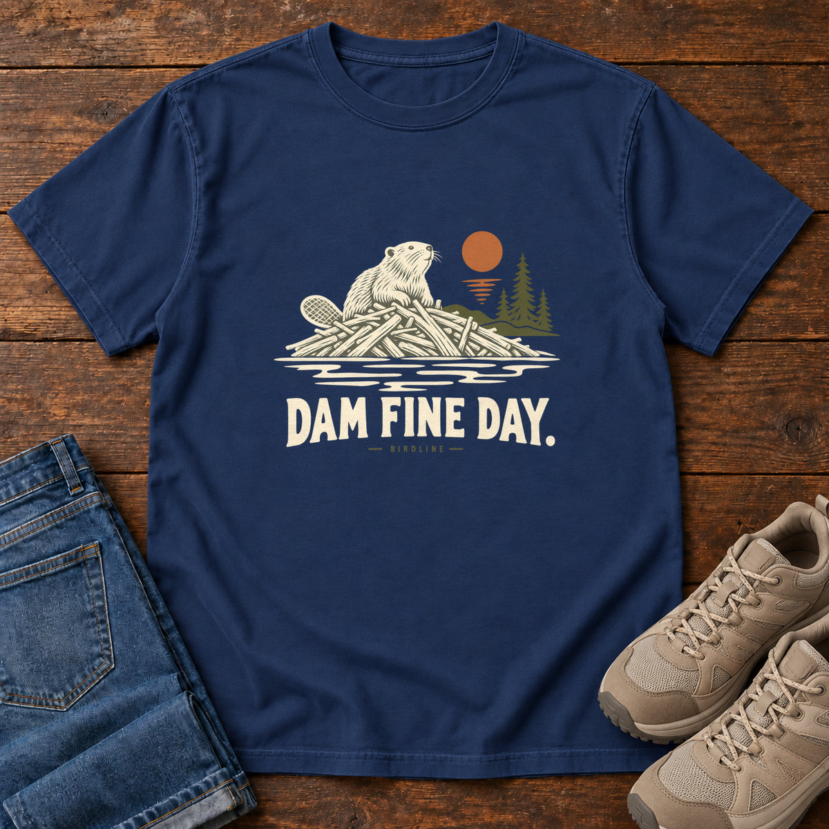 True Navy Dam Fine Day T-Shirt with a beaver on a pile of branches beside evergreens and an orange sun; front flat lay on wood.