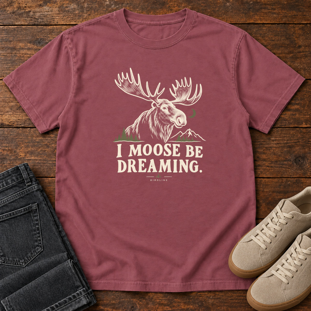 Brick I Moose Be Dreaming T-Shirt with a cream moose portrait with broad antlers, mountains and a crescent moon; front flat lay on wood.