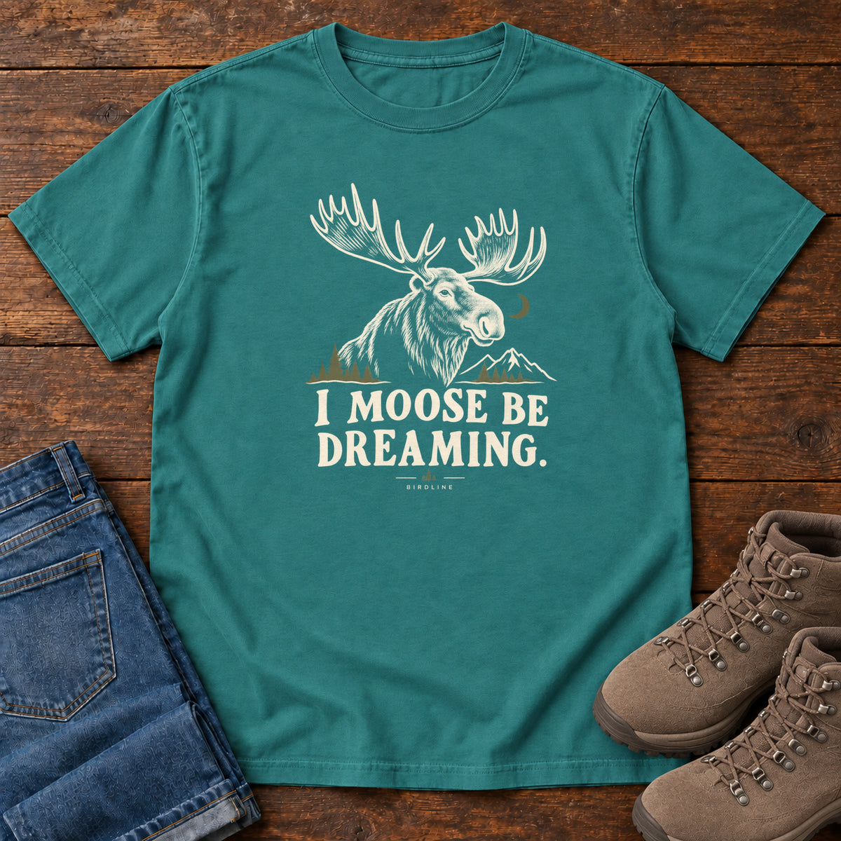 Emerald I Moose Be Dreaming T-Shirt with a cream moose portrait with broad antlers, mountains and a crescent moon; front flat lay on wood.