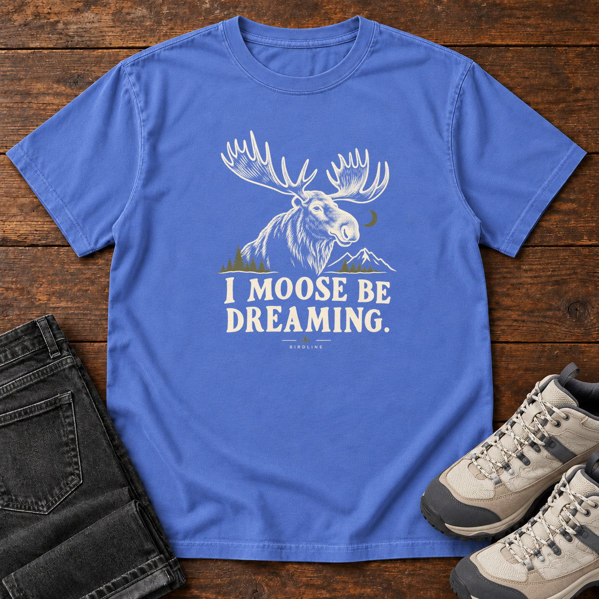 Mystic Blue I Moose Be Dreaming T-Shirt with a cream moose portrait with broad antlers, mountains and a crescent moon; front flat lay on wood.