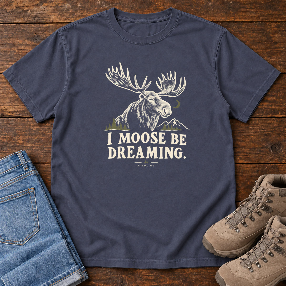 Navy I Moose Be Dreaming T-Shirt with a cream moose portrait with broad antlers, mountains and a crescent moon; front flat lay on wood.
