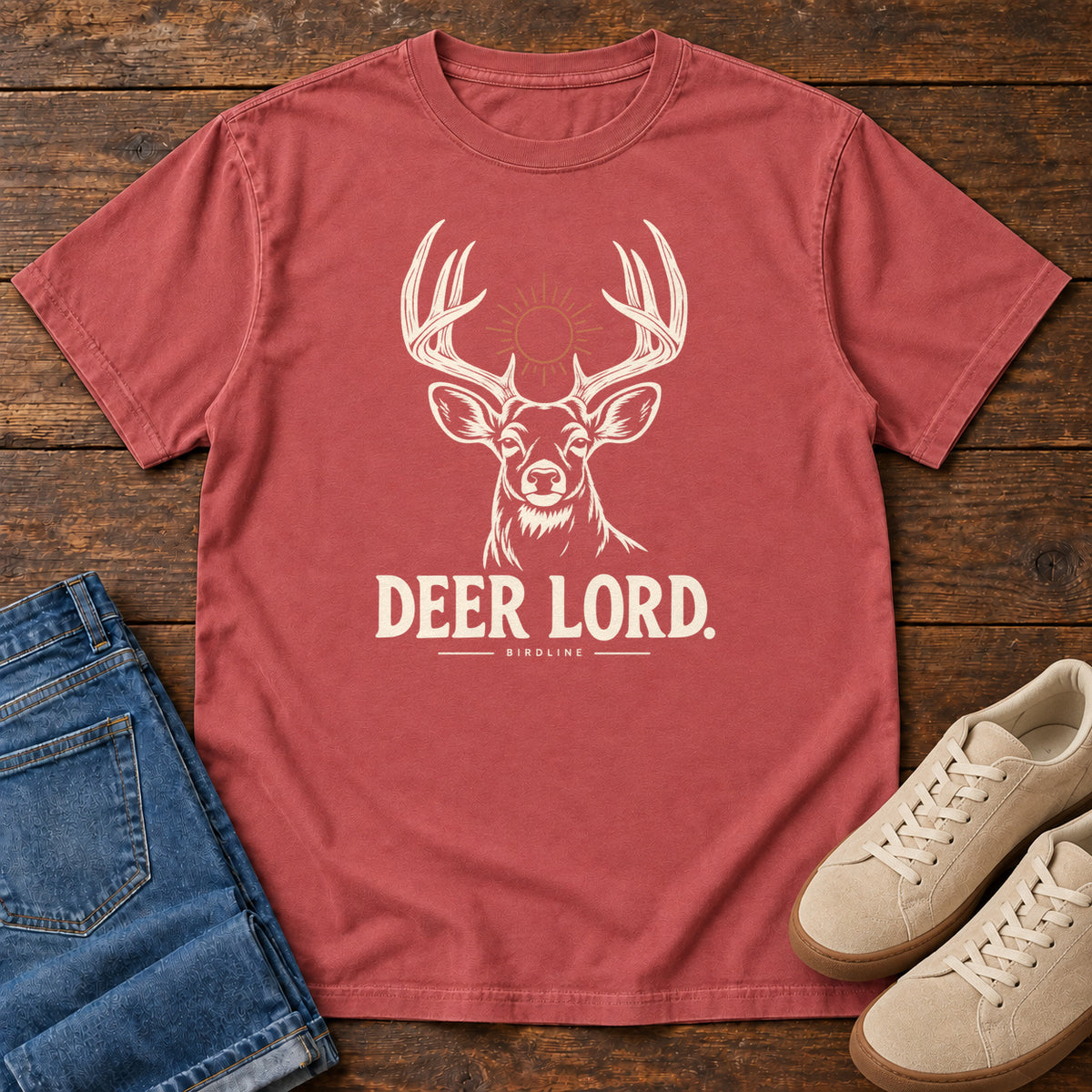 Crimson Deer Lord T-Shirt with a cream antlered deer head, a sun symbol and Deer Lord lettering; front flat lay on wood.