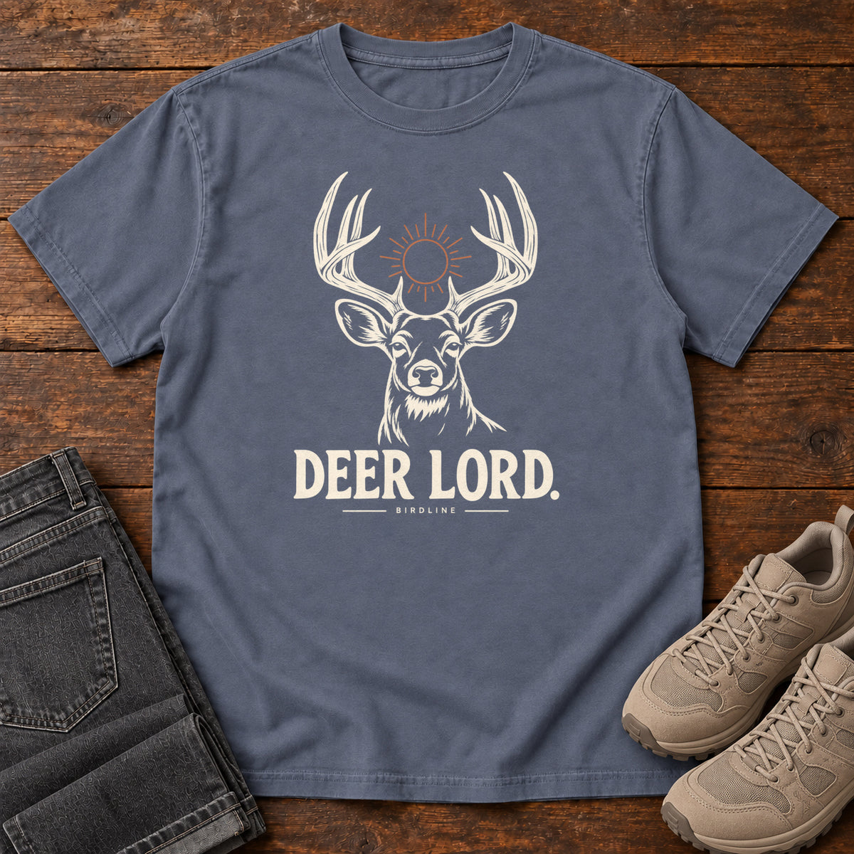 Denim Deer Lord T-Shirt with a cream antlered deer head, a sun symbol and Deer Lord lettering; front flat lay on wood.