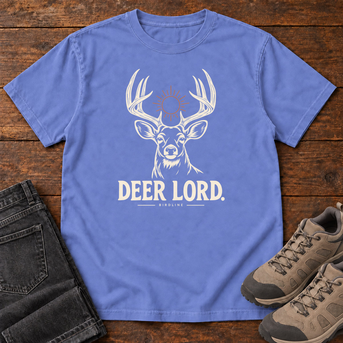 Flo Blue Deer Lord T-Shirt with a cream antlered deer head, a sun symbol and Deer Lord lettering; front flat lay on wood.