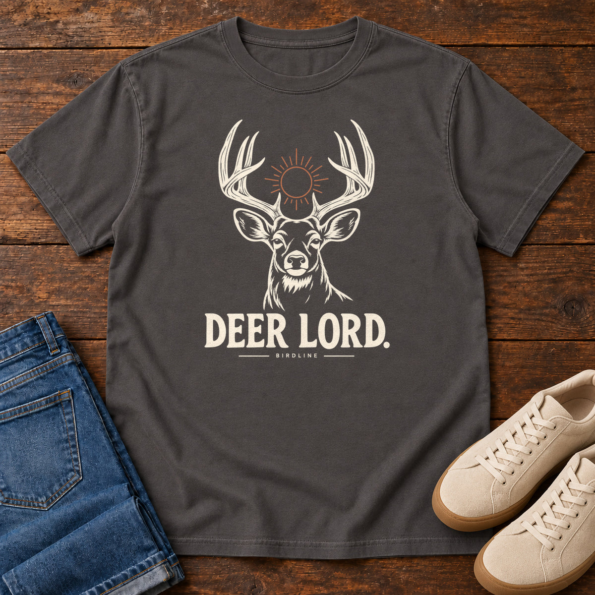 Graphite Deer Lord T-Shirt with a cream antlered deer head, a sun symbol and Deer Lord lettering; front flat lay on wood.