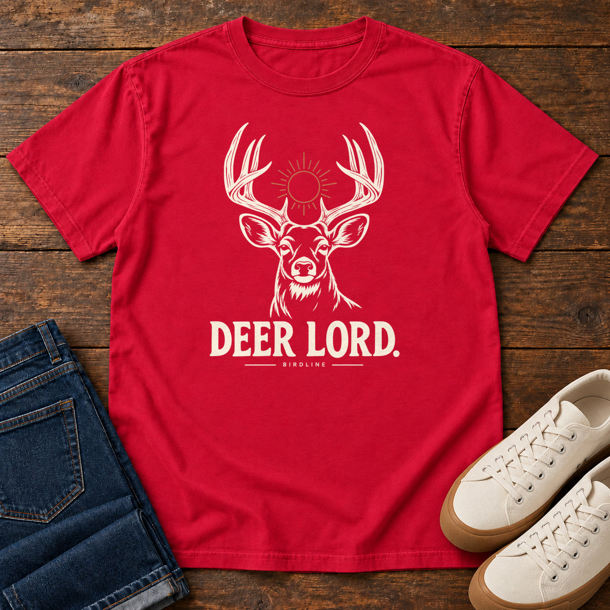 Red Deer Lord T-Shirt with a cream antlered deer head, a sun symbol and Deer Lord lettering; front flat lay on wood.