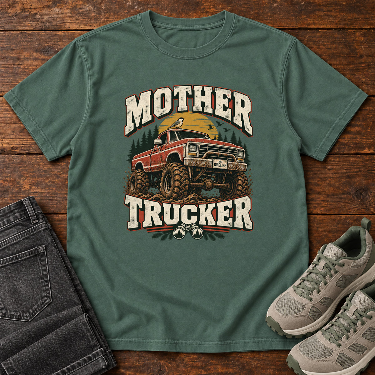 Blue Spruce Mother Trucker Red Truck T-Shirt with a lifted red pickup truck, evergreen trees and cream lettering, front flat lay on wood.