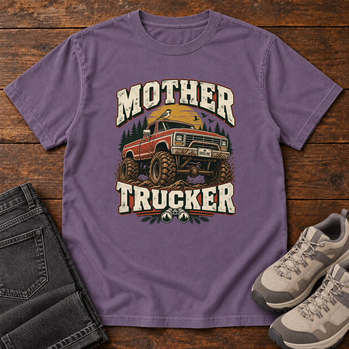 Grape Mother Trucker Red Truck T-Shirt with a lifted red pickup truck, evergreen trees and cream lettering, front flat lay on wood.