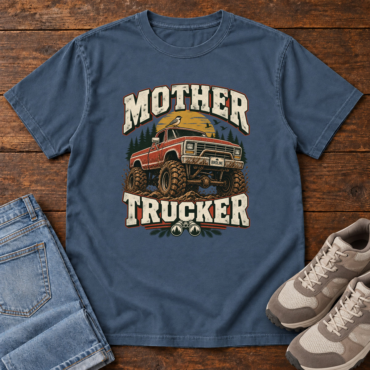 Midnight Mother Trucker Red Truck T-Shirt with a lifted red pickup truck, evergreen trees and cream lettering, front flat lay on wood.