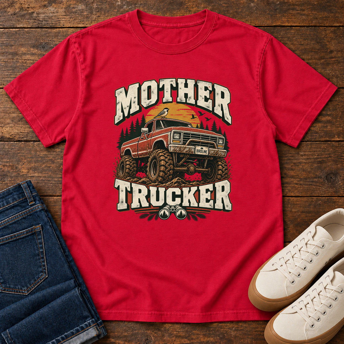 Red Mother Trucker Red Truck T-Shirt with a lifted red pickup truck, evergreen trees and cream lettering, front flat lay on wood.