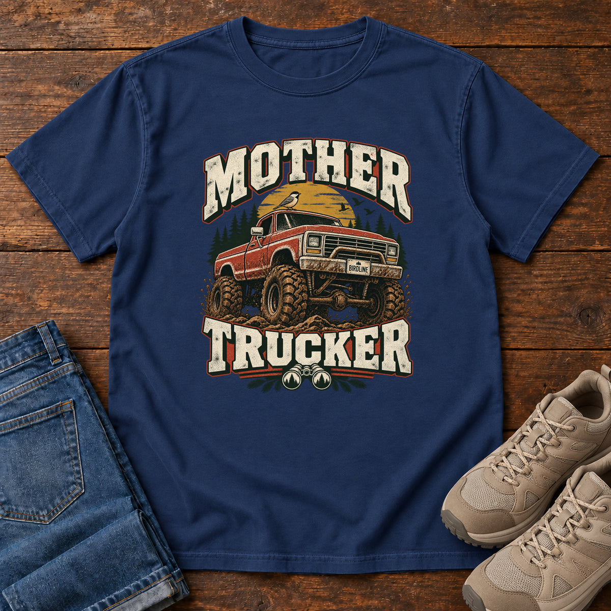 True Navy Mother Trucker Red Truck T-Shirt with a lifted red pickup truck, evergreen trees and cream lettering, front flat lay on wood.