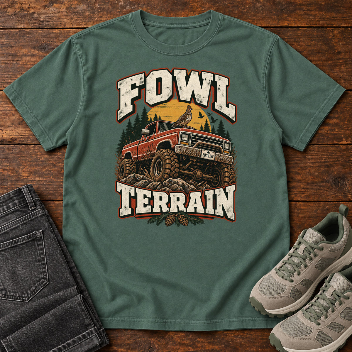 Fowl Terrain T-Shirt in Blue Spruce, front laid flat on wood, featuring an orange off-road truck with a bird on its roof, pine trees, a yellow sun and bold cream lettering.