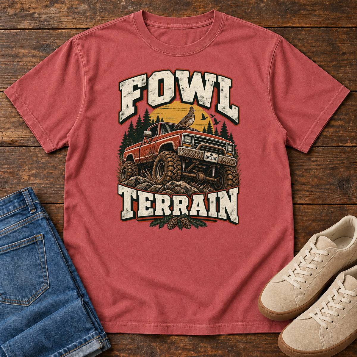 Fowl Terrain T-Shirt in Crimson, front laid flat on wood, featuring an orange off-road truck with a bird on its roof, pine trees, a yellow sun and bold cream lettering.
