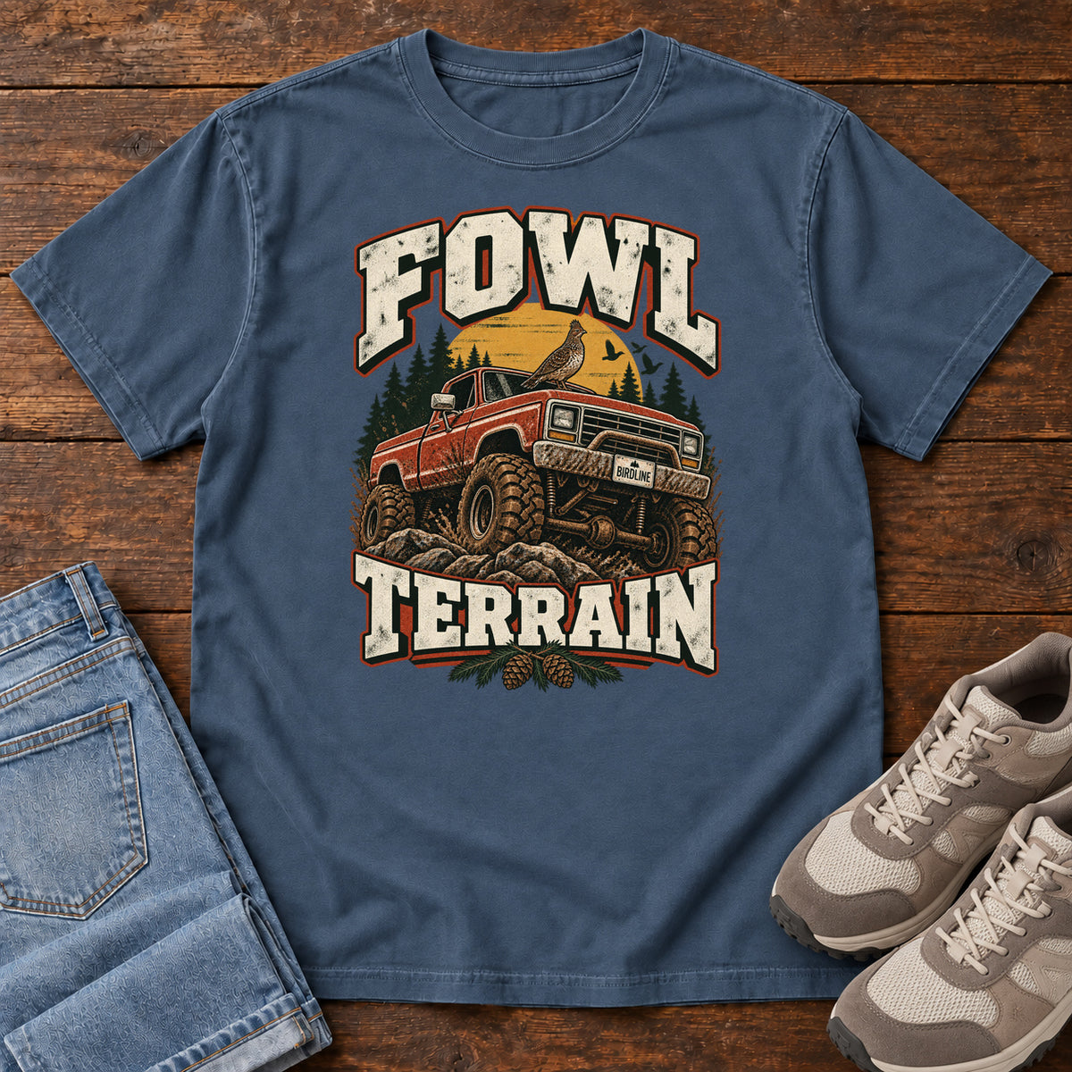 Fowl Terrain T-Shirt in Midnight, front laid flat on wood, featuring an orange off-road truck with a bird on its roof, pine trees, a yellow sun and bold cream lettering.