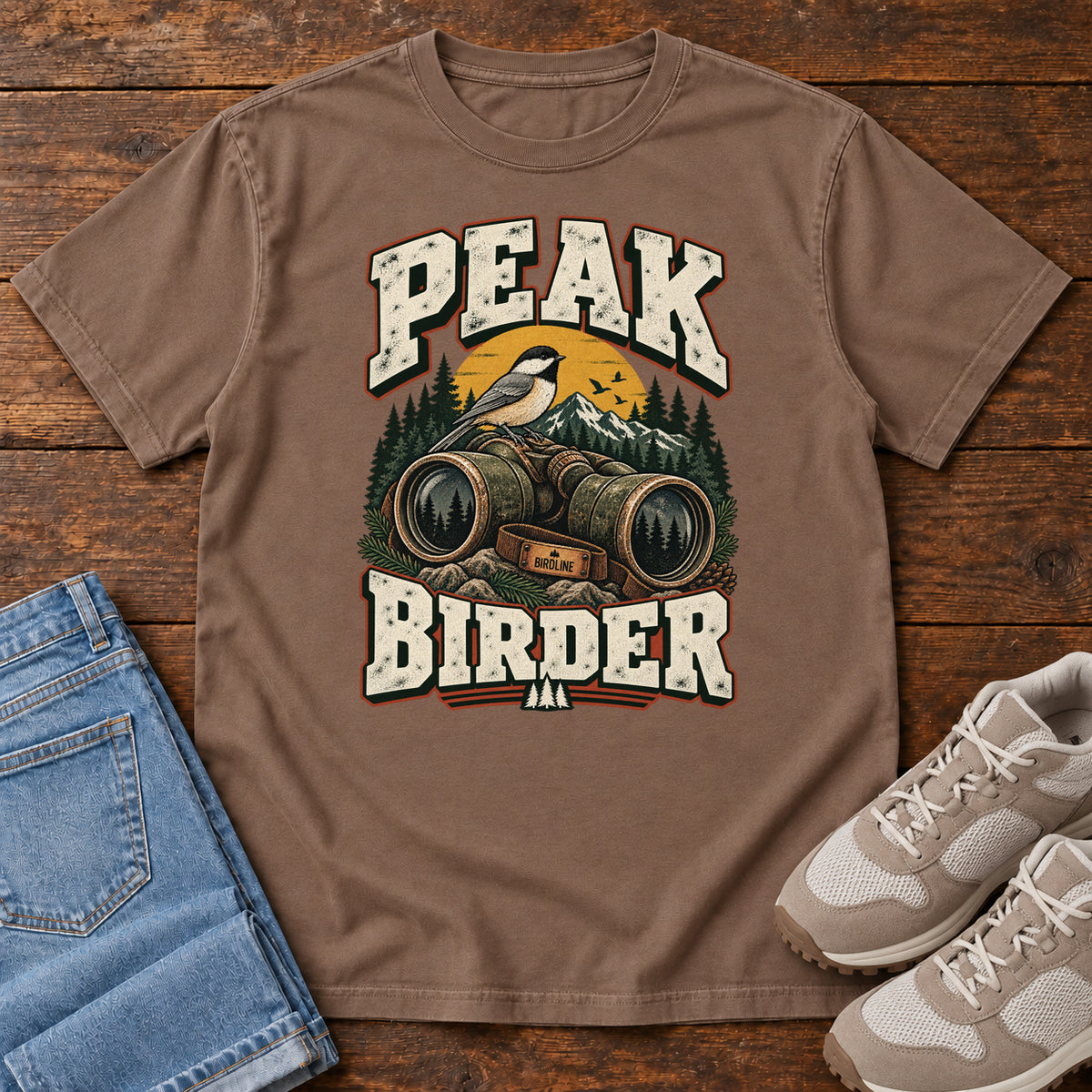 Espresso Peak Birder T-Shirt with a small black-capped bird on binoculars, with mountains and distressed cream lettering, front flat lay on wood.