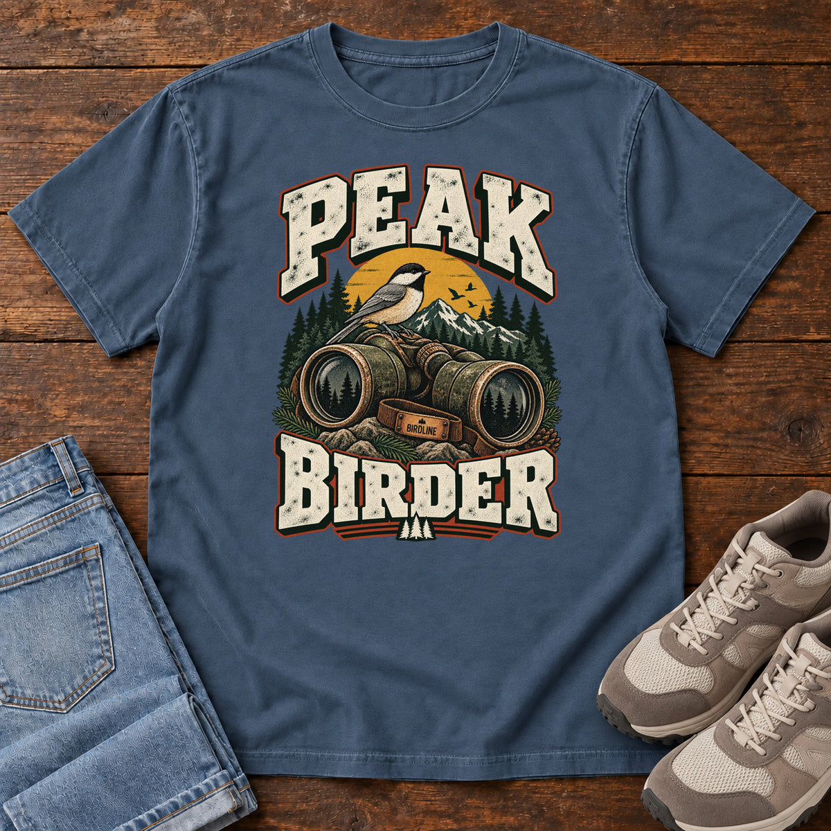Midnight Peak Birder T-Shirt with a small black-capped bird on binoculars, with mountains and distressed cream lettering, front flat lay on wood.