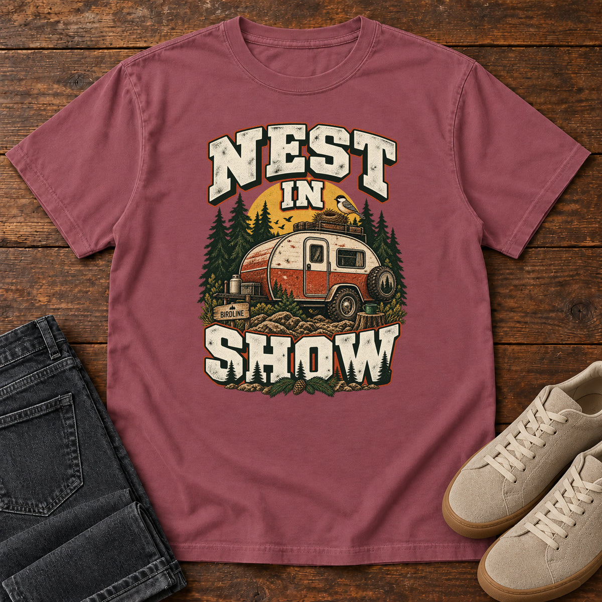 Brick Nest in Show T-Shirt with a red-and-cream camper beneath pines, with a small bird and nest on its roof; front flat lay on wood.