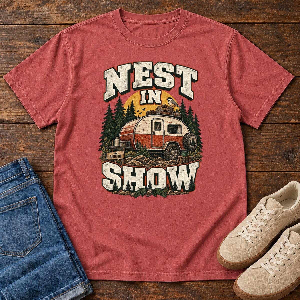 Crimson Nest in Show T-Shirt with a red-and-cream camper beneath pines, with a small bird and nest on its roof; front flat lay on wood.
