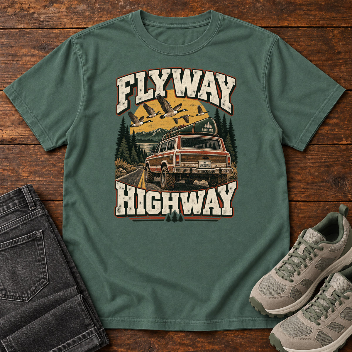 Blue Spruce Flyway Highway T-Shirt with a vintage pickup on a forest road with flying birds overhead; front flat lay on wood.