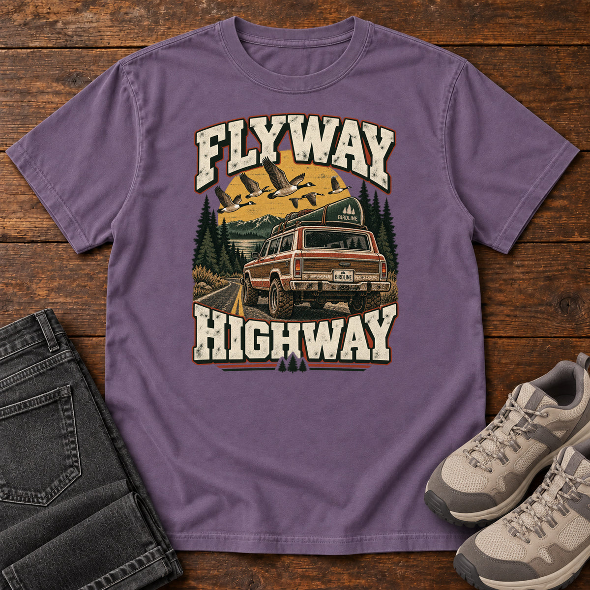 Grape Flyway Highway T-Shirt with a vintage pickup on a forest road with flying birds overhead; front flat lay on wood.