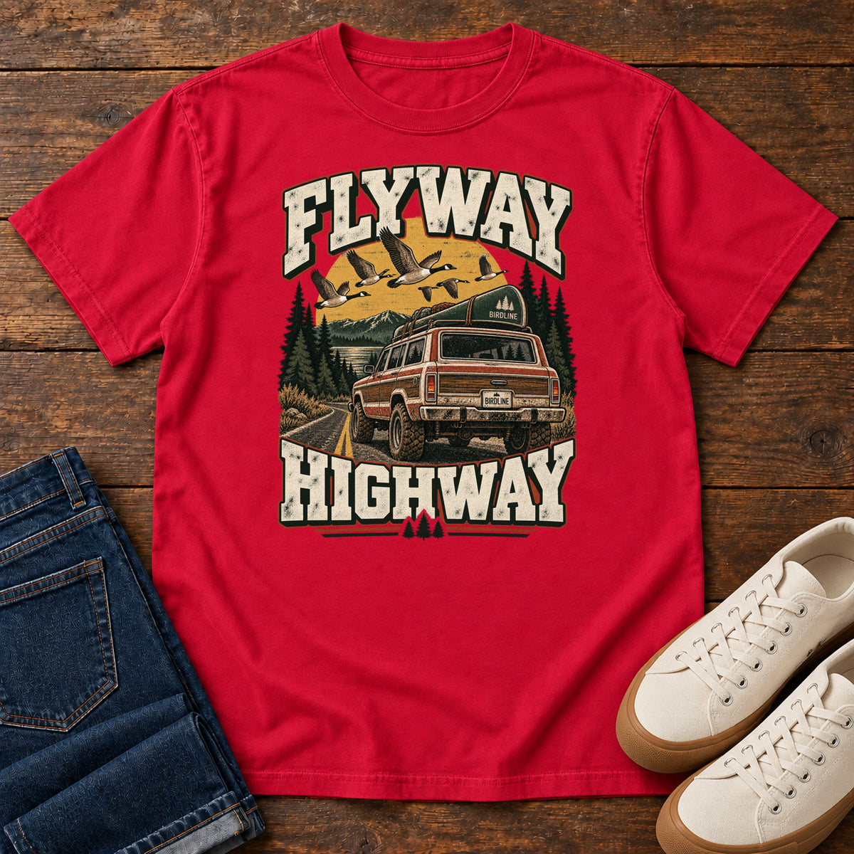 Red Flyway Highway T-Shirt with a vintage pickup on a forest road with flying birds overhead; front flat lay on wood.