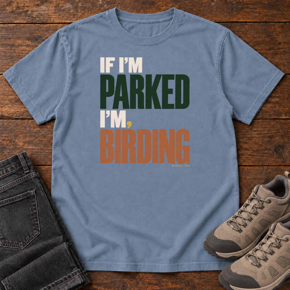 If I'm Parked I'm Birding T-Shirt in Blue Jean, front laid flat on wood, featuring stacked cream, dark green and rust slogan lettering with a small yellow dot.