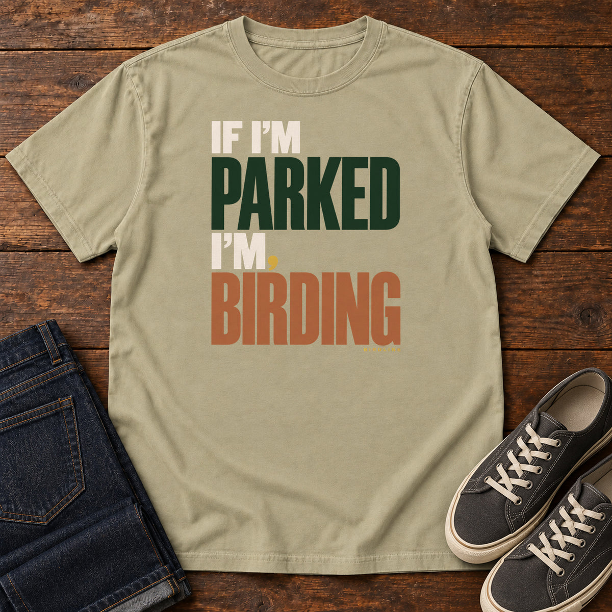If I'm Parked I'm Birding T-Shirt in Khaki, front laid flat on wood, featuring stacked cream, dark green and rust slogan lettering with a small yellow dot.